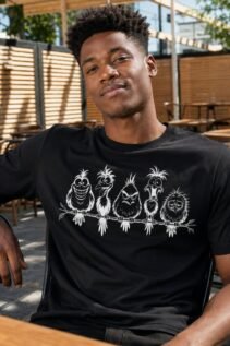 Mann trägt schwarzes T-Shirt mit weißem frechen Tiermotiv aus Vögeln im Café, sitzende Frontansicht in entspannter Pose.