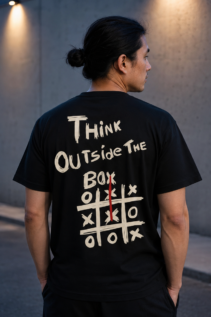 Rückansicht eines Mannes im schwarzen T-Shirt mit großem „Think Outside the Box“-Aufdruck, draußen vor einer grauen Betonwand.
