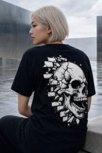 Schwarzes Skull T-Shirt an einer sitzenden Frau mit blondem Bob, Rückenansicht in moderner Betonkulisse, großes weißes Totenkopf-Motiv sichtbar.