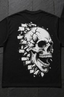 Schwarzes T-Shirt flach auf dunklem Steinboden ausgelegt, großes weißes Skull-Motiv auf der Rückseite, kleines Motiv auf einer Karte daneben.