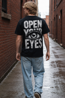 Mann trägt schwarzes Open Your Eyes T-Shirt von hinten in einer nassen Backstein-Gasse, sichtbares Auge-Motiv und streetwear shirt Stil.