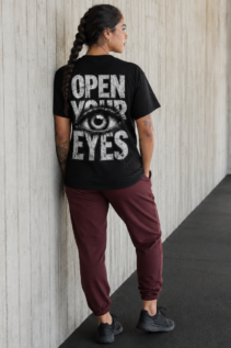 Frau trägt schwarzes Open Your Eyes T-Shirt von hinten an heller Betonwand, schwarzes Auge-Motiv sichtbar, lässige alternative mode mit Streetwear-Anmutung.
