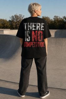 Kurzhaarige Person von hinten in schwarzem Statement T-Shirt mit rotem und weißem Aufdruck „THERE IS NO COMPETITION.“ auf einer Skatepark-Rampe.