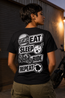 Frau von hinten trägt ein schwarzes Eat Sleep Ride T-Shirt vor einer Garage bei Sonnenuntergang, daneben steht ein Motorrad, motivischer Statement-Shirt-Look