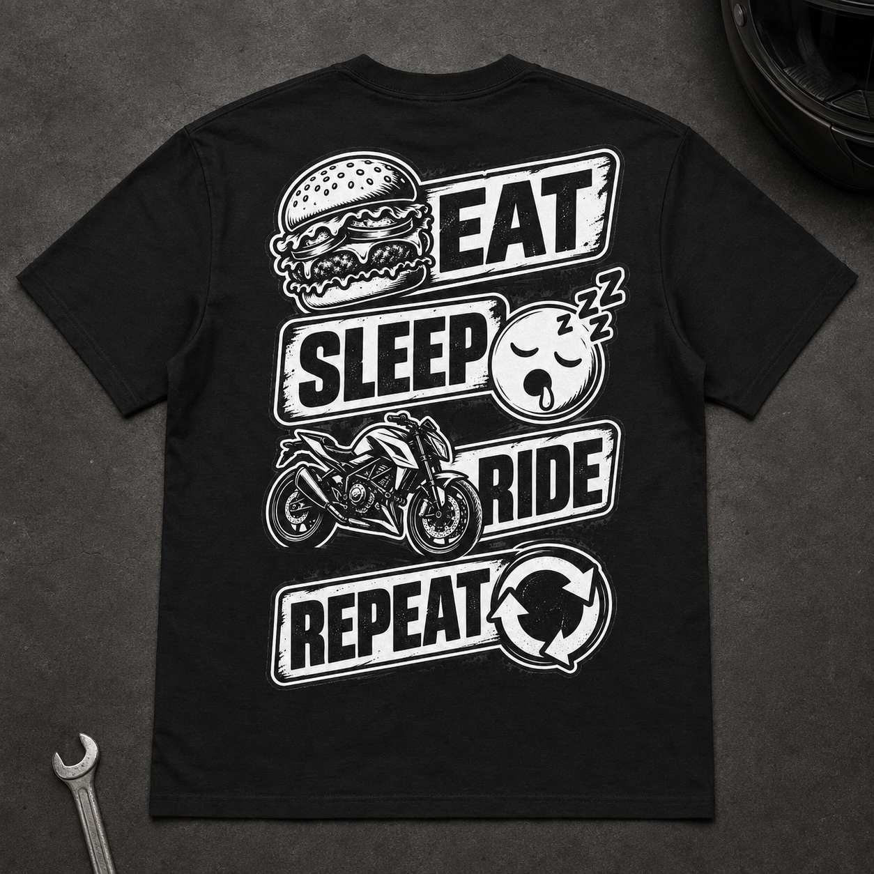 Schwarzes T-Shirt flach ausgelegt mit großem Eat Sleep Ride Rückendruck, neben einem Schraubenschlüssel auf dunkler Werkbank