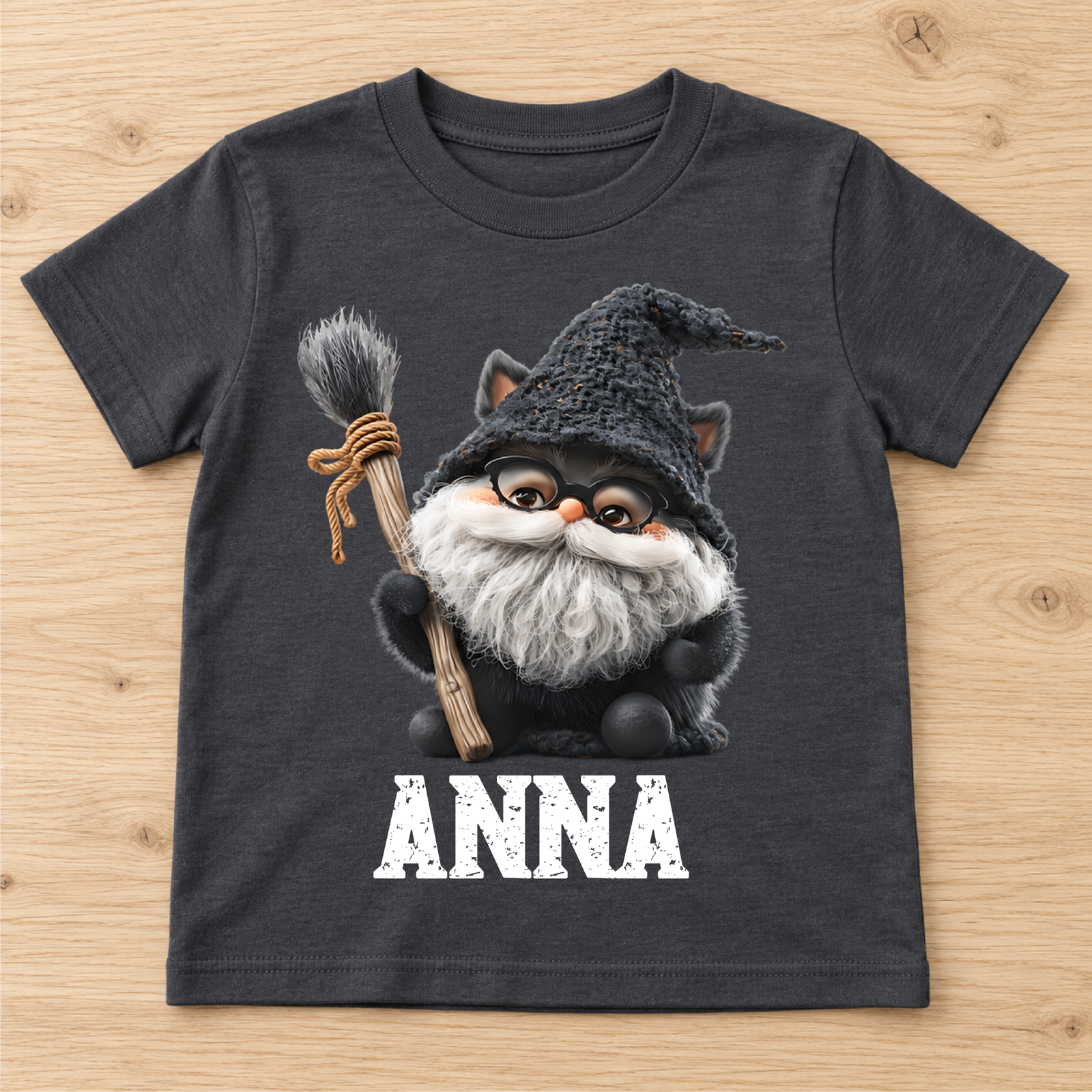Dunkelgraues Kinder-T-Shirt mit Hexenkatze-Motiv und dem Namen ANNA liegt flach auf hellem Holzhintergrund
