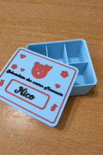 Offene personalisierte Schatzkiste aus 3D-Druck in Hellblau mit rotem Deckelmotiv Nico, schräg auf Holztisch mit sichtbarem Innenfach.