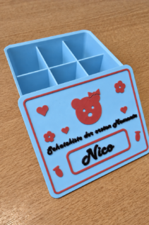 Schräg geöffnete 3D-gedruckte Schatzkiste in Hellblau mit rotem Teddy-Motiv und Name Nico, auf Holztisch mit offenem Fach-Innenleben.