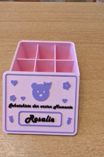 Freigestellte 3D-gedruckte personalisierte Schatzkiste in Pastellrosa und Lila mit offenem Deckel, innen mit Fächern, auf Holztisch mit Name Rosalie und Teddy-Motiv.